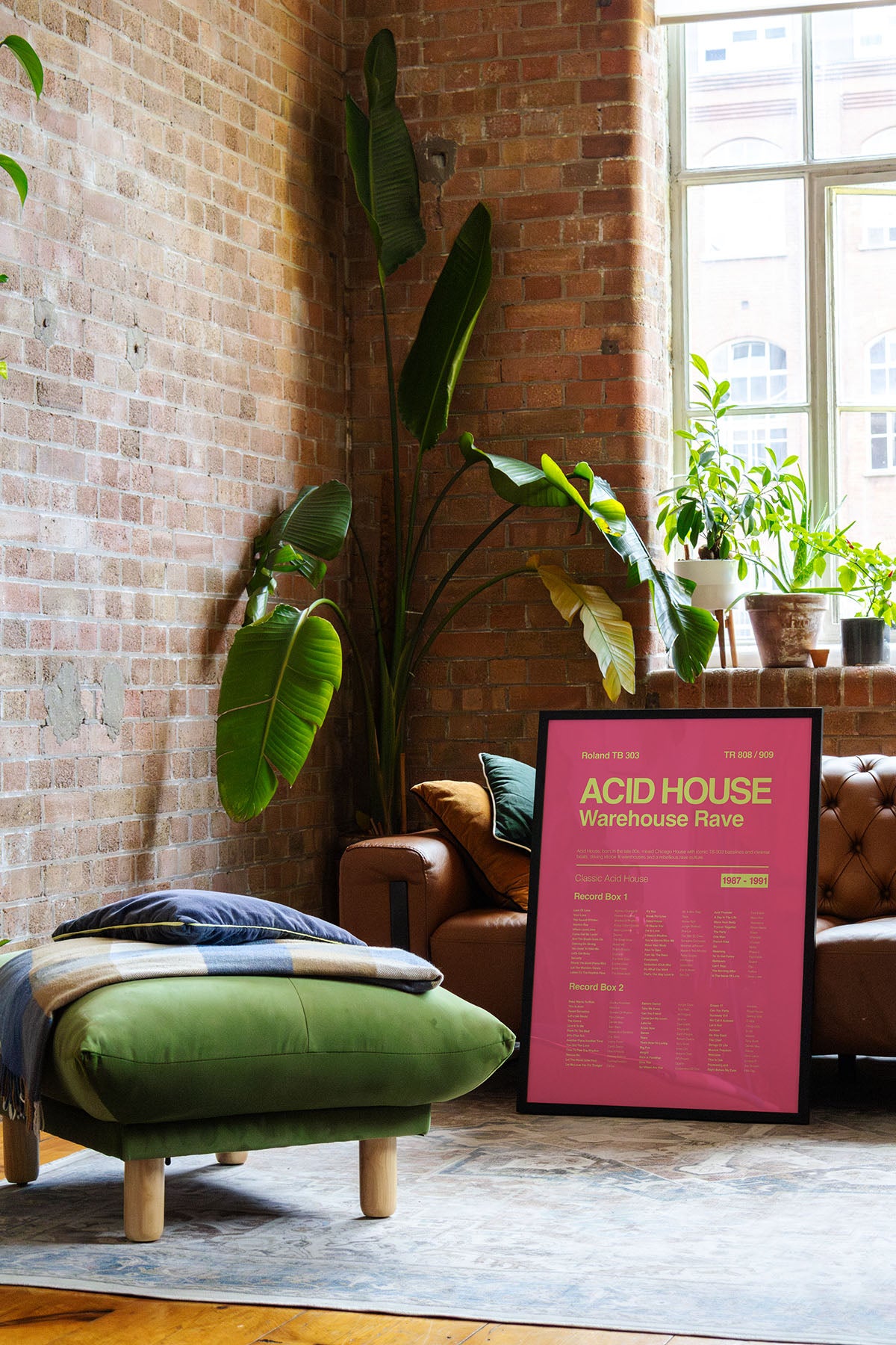 Acid House Rave music print in a black frame resting on a brown leather sofa in a bright, modern loft with exposed brick walls and natural reflections. Suitable for A1, A2 and A3 sizes. Electric pink background with acid yellow text (Mockup M12).