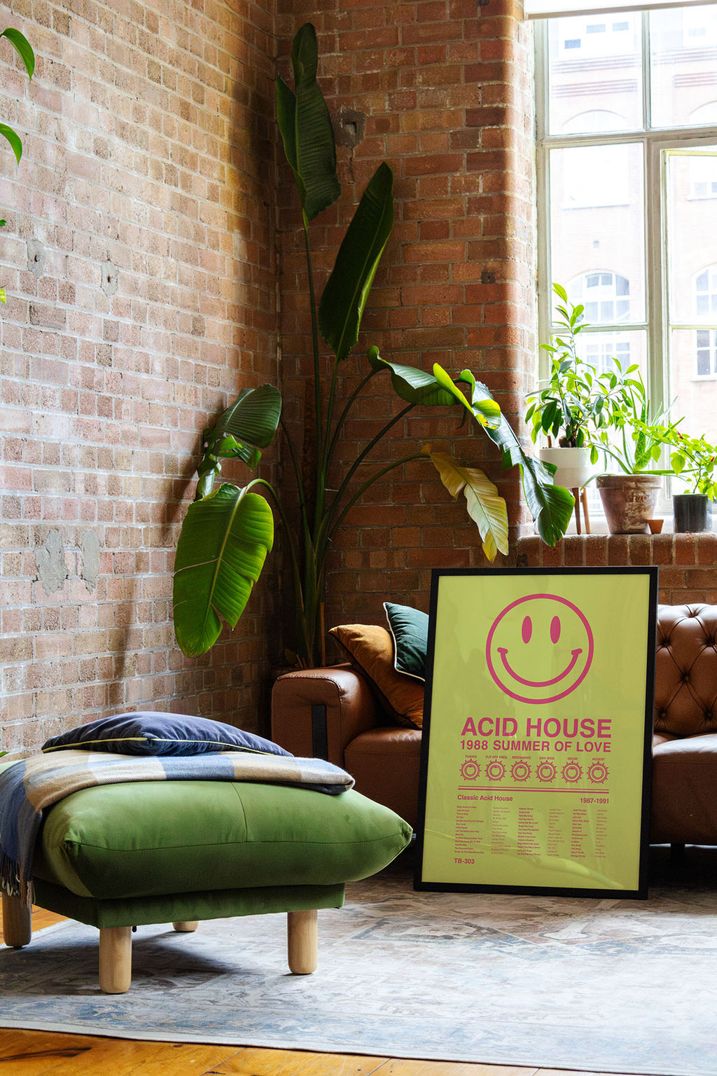 Acid House Smiley Face music print in a black frame leaning against a brown leather sofa in a bright exposed brick loft apartment, with natural window reflections across the frame. Suitable for A1, A2 and A3 sizes. Acid yellow background with electric pink text (Mockup M12).