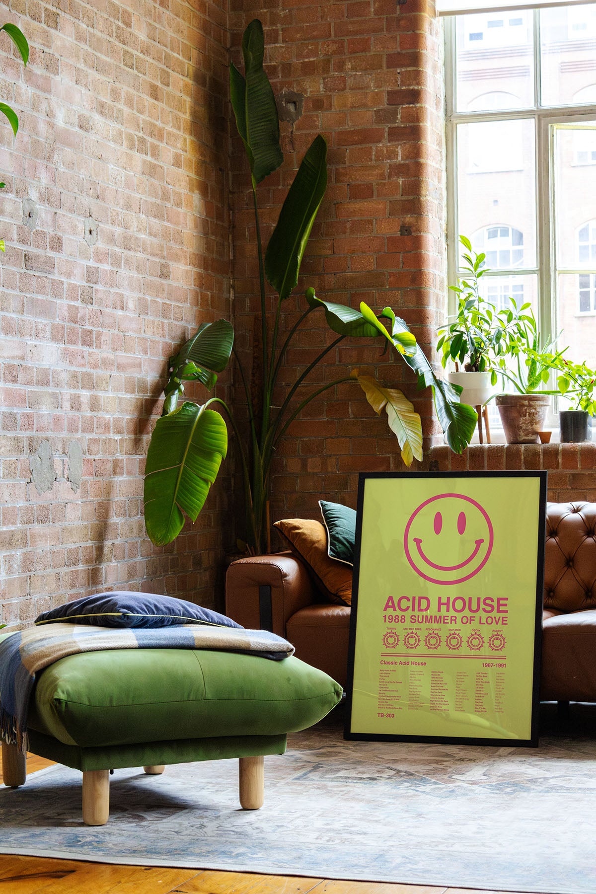 Acid House Smiley Face music print in a black frame leaning against a brown leather sofa in a bright exposed brick loft apartment, with natural window reflections across the frame. Suitable for A1, A2 and A3 sizes. Acid yellow background with electric pink text (Mockup M12).