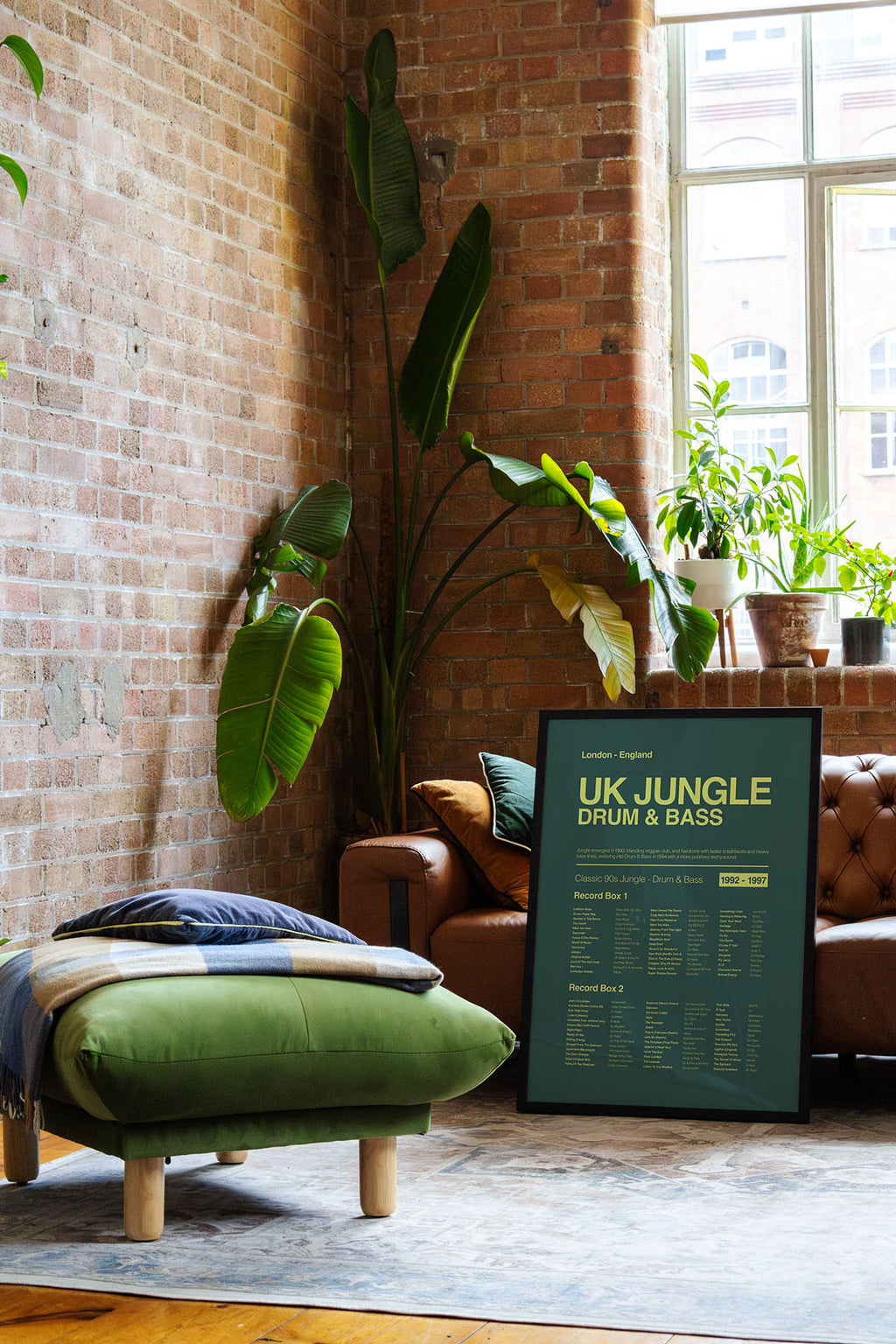 Jungle Drum and Bass music print in a black frame leaned against a leather sofa in a bright loft with exposed brick walls and natural window reflections. Suitable for A1, A2 and A3 sizes. Green background with acid yellow text (Mockup M12).