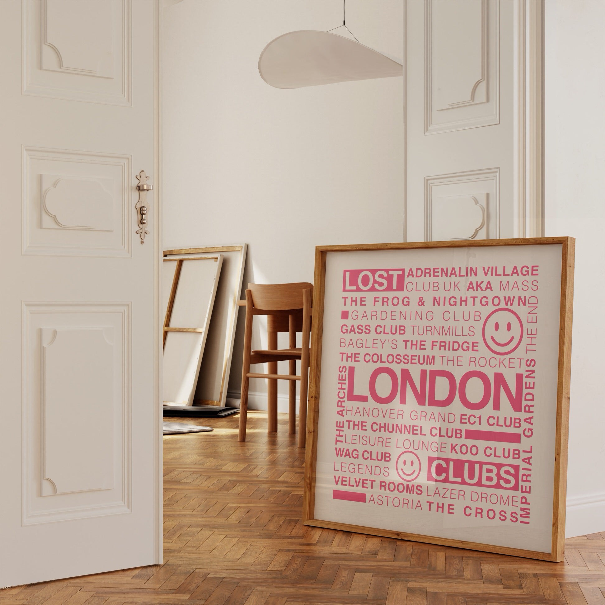 Lost London Clubs square music print with electric pink text on white in a wooden frame leaning against a white ornate wall in a doorway